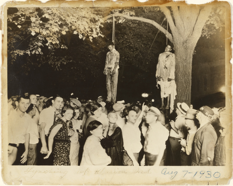 Black and white photograph of two black men hung from a tree while smiling and laughing white people celebrate.