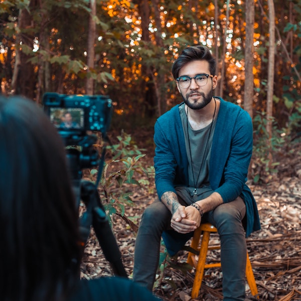 A
    smiling man, sitting sitting in the forest in front of a camera.