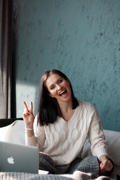 A smiling woman, sitting on a bed with her computer, flashin a peace sign.